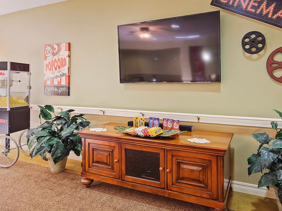A cozy entertainment area with a large flat-screen TV mounted on a light green wall above a wooden cabinet. On the cabinet are various snack boxes and napkins. To the left, there is a popcorn machine and a potted plant. The wall is decorated with a vintage popcorn sign and film reel decorations.