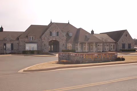 Exterior view of a large stone building with multiple peaked roofs and windows, featuring a stone sign in front that reads Heritage Place. The building is set against a cloudy sky with a paved road and parking area in the foreground.