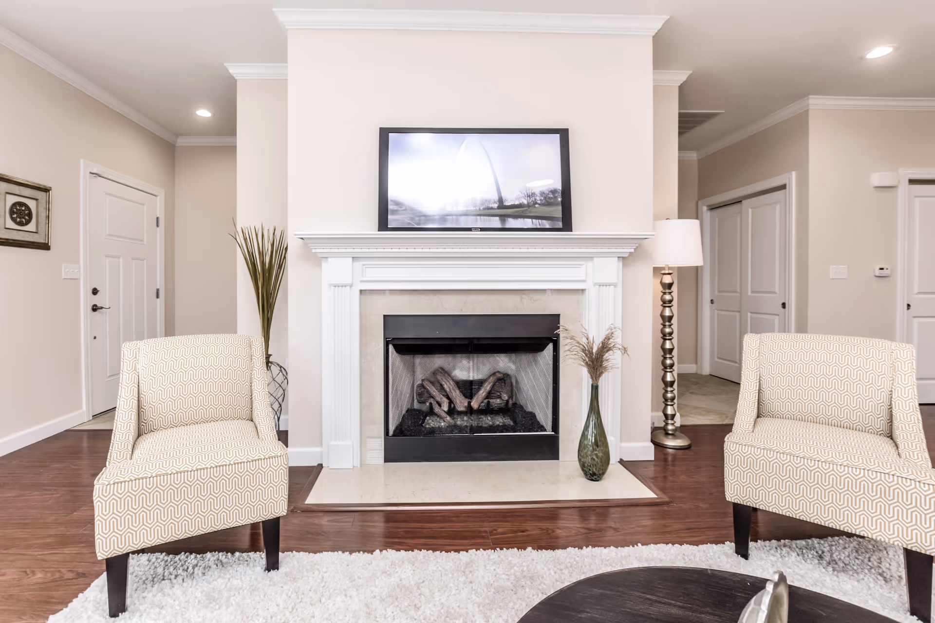 A bright living room featuring a white fireplace with a framed picture above it, two patterned armchairs on either side, a tall floor lamp, a vase with dried plants, and hardwood flooring with a white rug.