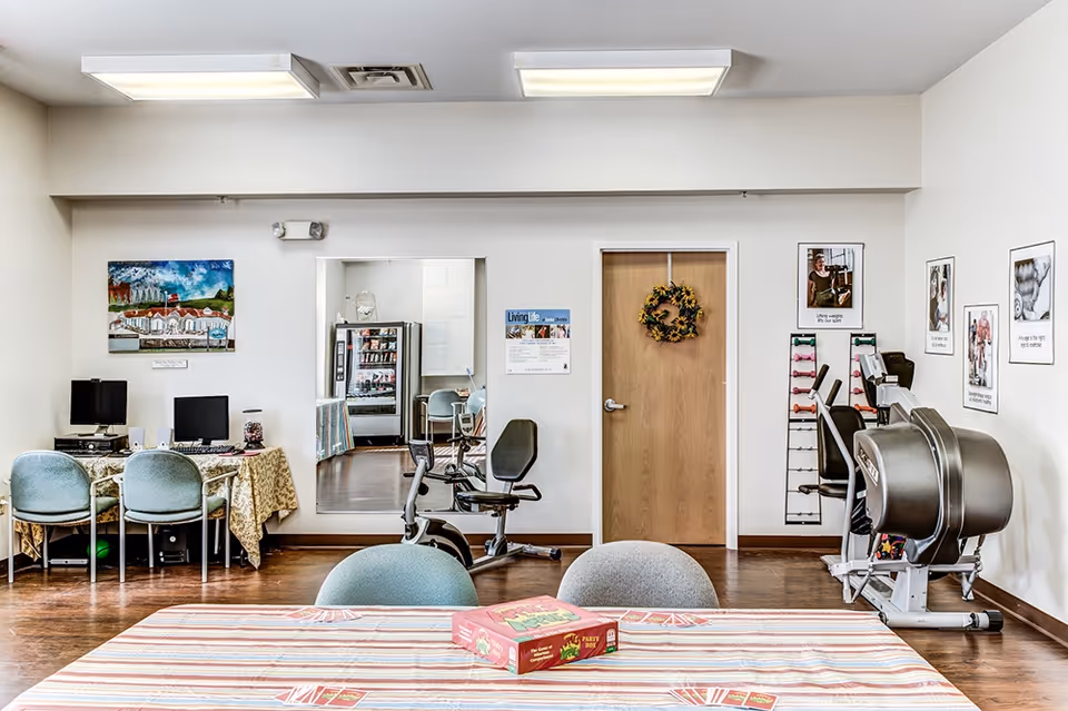 Bright multipurpose activity room with tables, chairs, computers and exercise equipment in a senior living facility.
