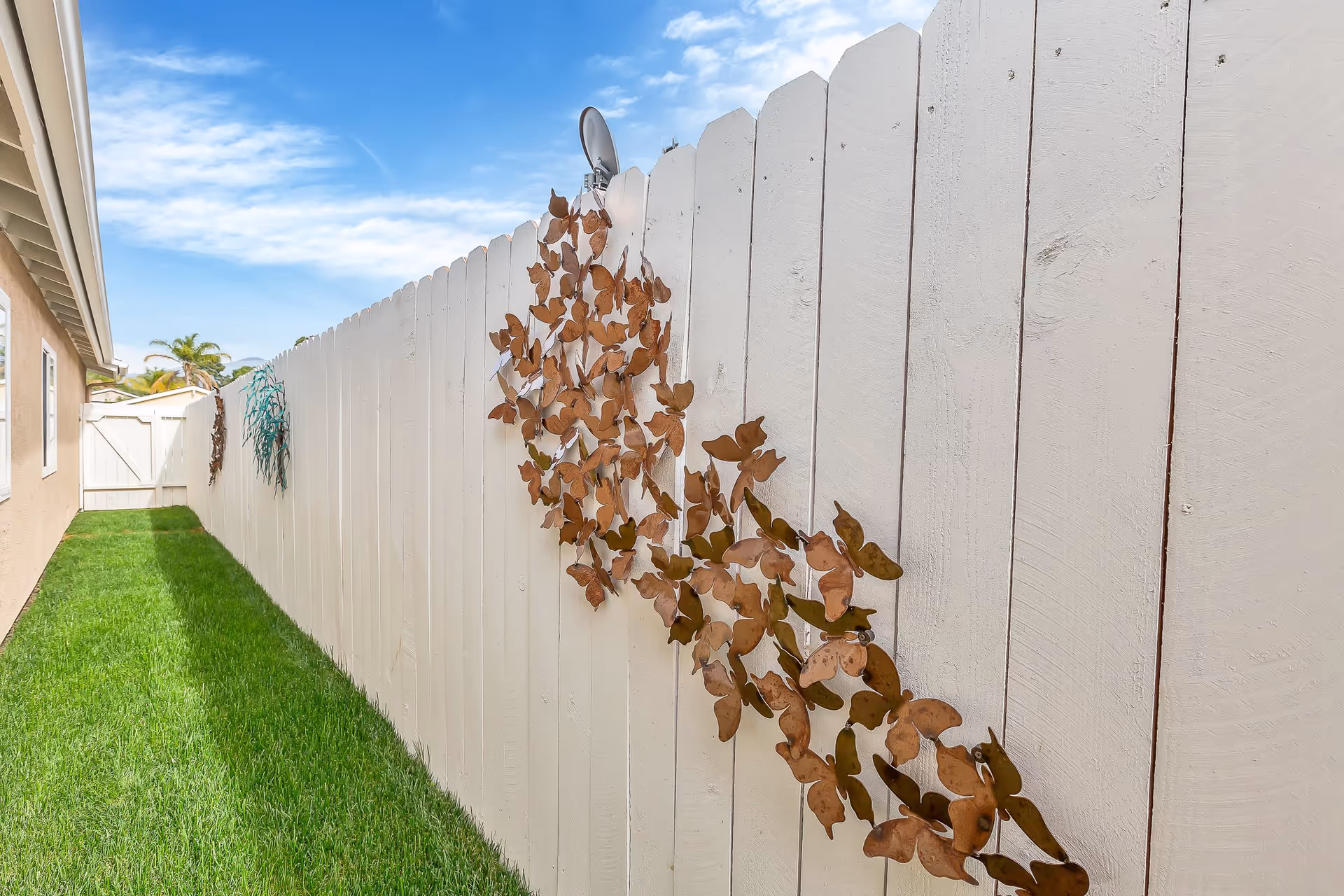 Narrow grassy side yard with a white wooden fence adorned with metal butterfly decorations under a blue sky.