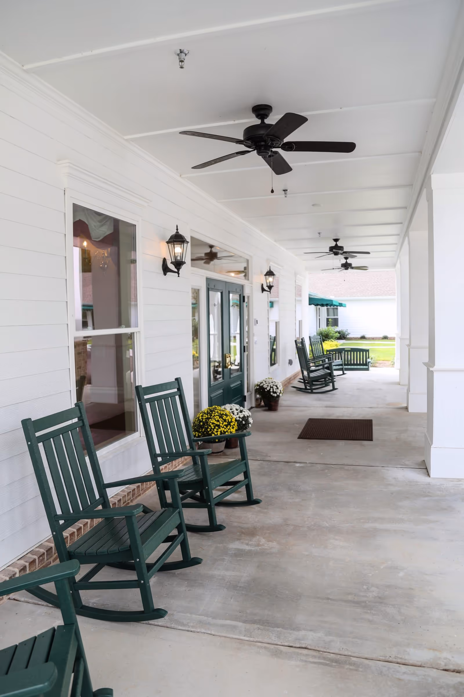 A covered outdoor porch area with several green wooden rocking chairs lined up along a white wall with windows and green double doors. There are ceiling fans mounted on the white ceiling and wall-mounted lantern-style lights. Potted flowers are placed near the doors, and a brown doormat is on the concrete floor. The porch overlooks a grassy area with shrubs and another building in the background.