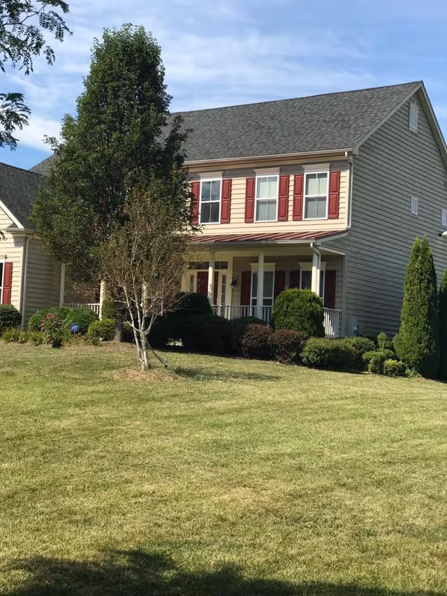 A two-story residential building with beige siding and red shutters on the windows. The house has a covered front porch with white pillars and is surrounded by neatly trimmed bushes and a green lawn. A tree stands in the front yard under a partly cloudy sky.