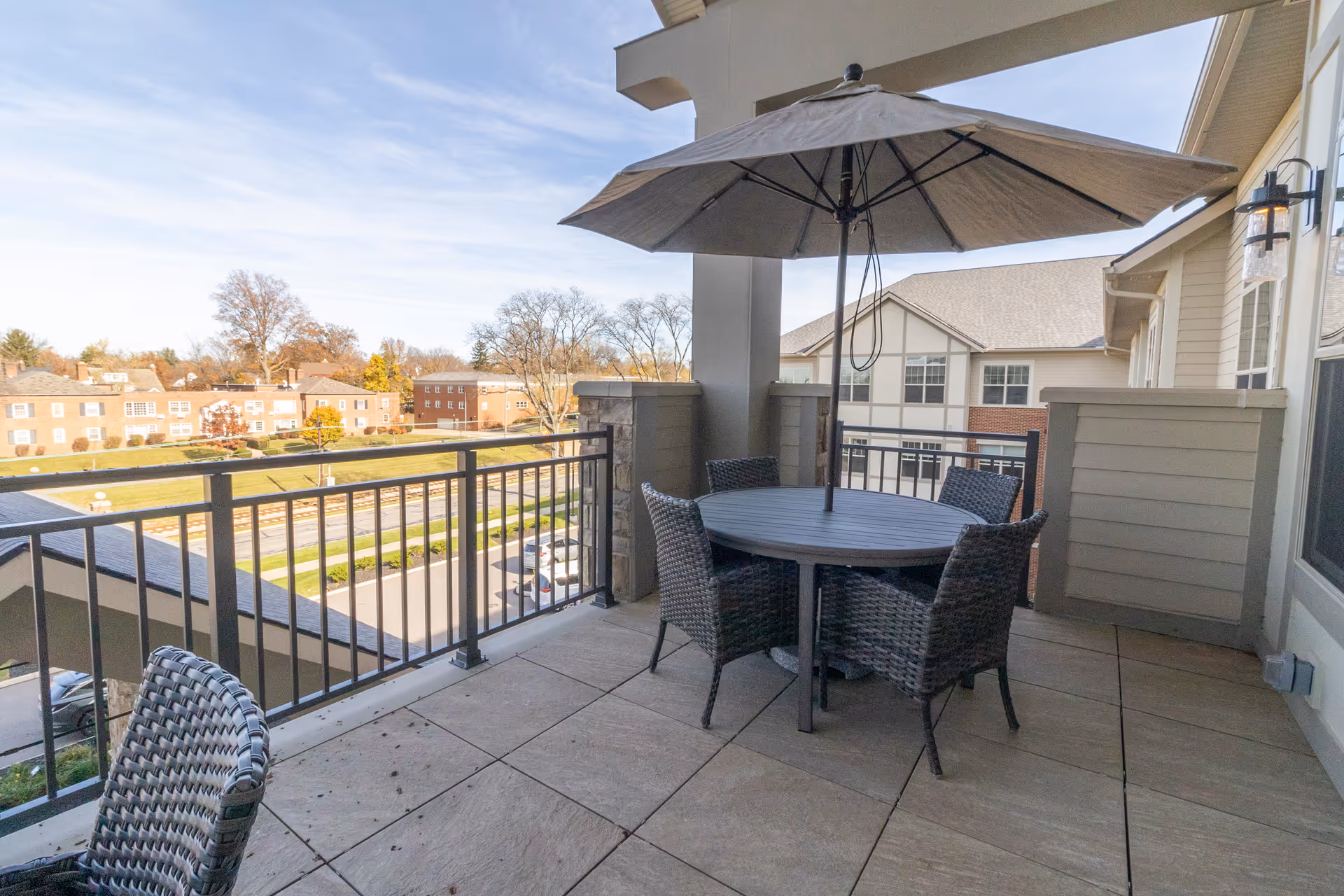 A covered balcony with a round table, umbrella and four wicker chairs overlooking nearby buildings and a parking area.