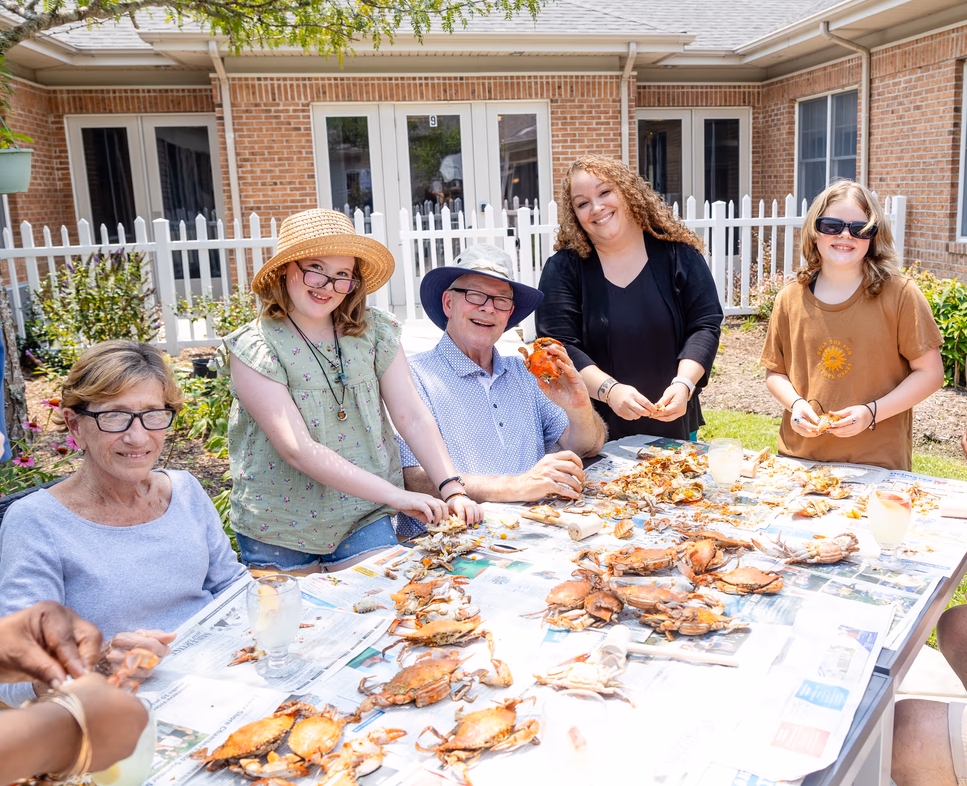 A group of five people, including seniors and younger individuals, sitting around a table outdoors enjoying a crab feast. The table is covered with newspapers and numerous cooked crabs. They are smiling and appear to be having a good time in a courtyard area with brick buildings and white picket fencing in the background.