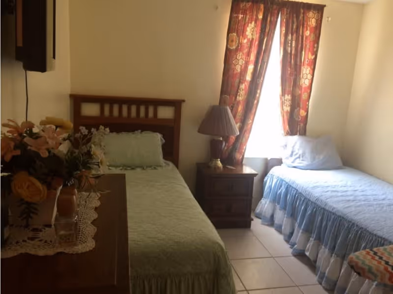 A small bedroom with two single beds, one with a green bedspread and the other with a blue bedspread. There is a wooden nightstand with a lamp between the beds, a window with patterned red curtains behind the nightstand, and a dresser with a floral arrangement on top to the left.