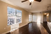 Empty room with large windows letting in natural light, dark wood flooring, beige walls, a ceiling fan with light fixture, and an open layout leading to another area of the home.