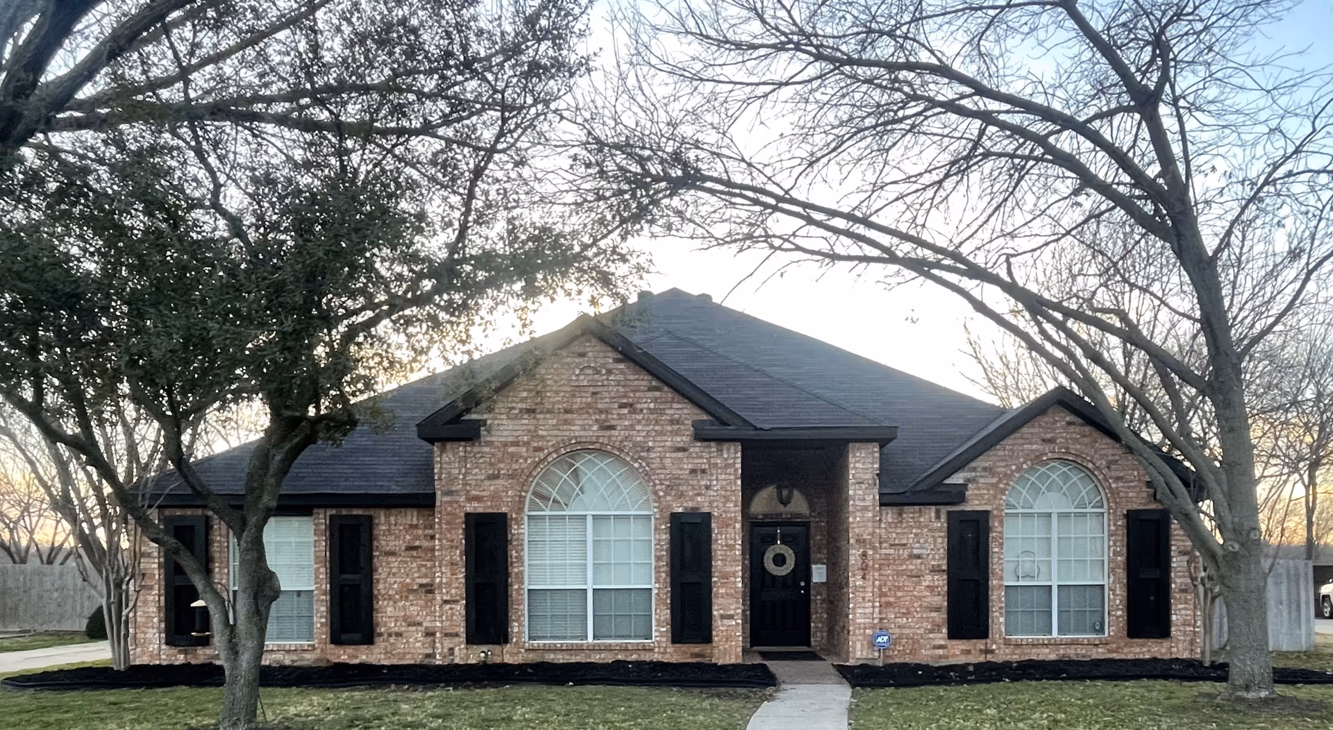 Single-story brick house front with large arched windows, a central black door with a wreath, and trees in the yard.