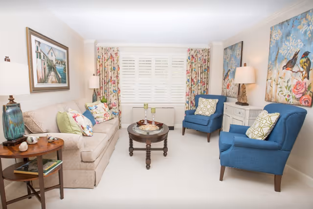 Bright furnished living room with a beige sofa, two blue armchairs, a round coffee table, and floral curtains over shuttered windows.