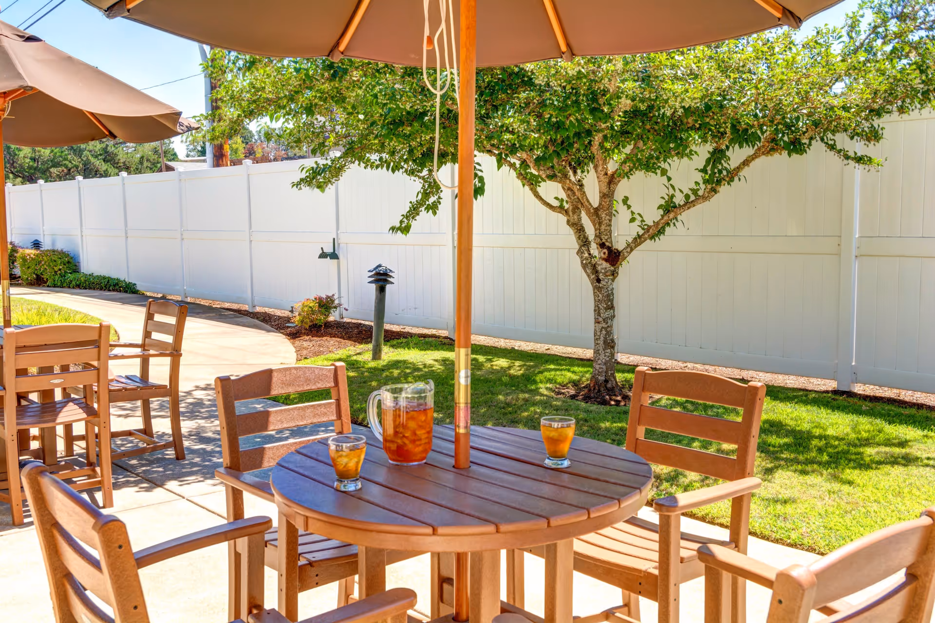 Outdoor patio area with wooden tables and chairs under large beige umbrellas. On one table, there is a pitcher and two glasses filled with iced tea. The patio is surrounded by a white fence, green grass, and a tree providing shade.