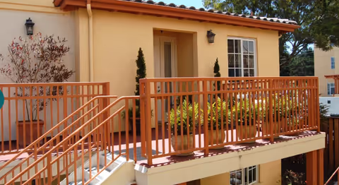 Exterior view of a senior living facility showing a beige building with a red railing balcony and stairs. There are potted plants on the balcony and two wall-mounted lantern-style lights near the entrance door. Trees and another building are visible in the background.