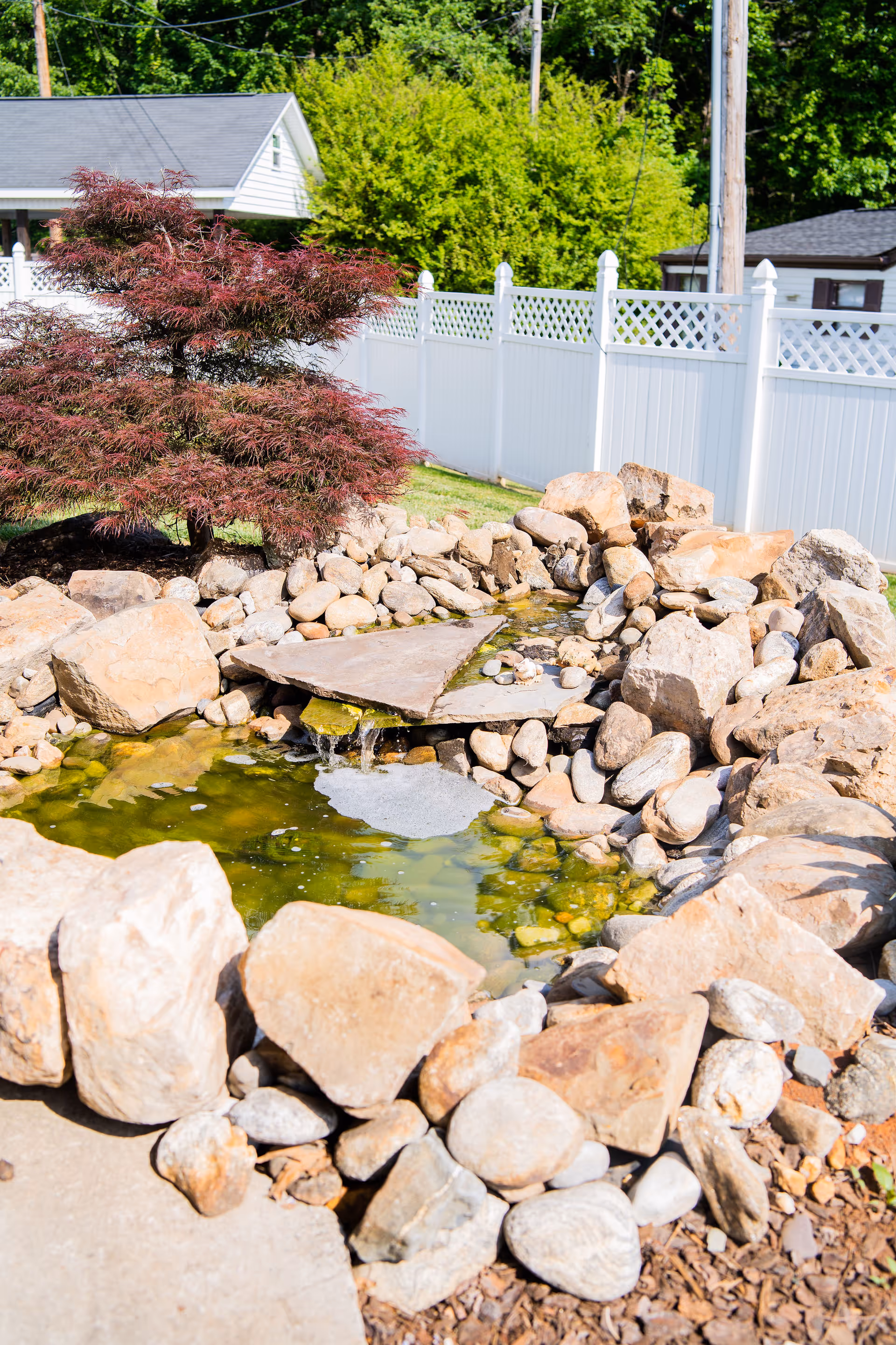 A small outdoor water feature surrounded by large rocks and pebbles, with a small waterfall flowing into a pond. There is a red-leafed tree to the left and a white fence in the background with green trees beyond it.