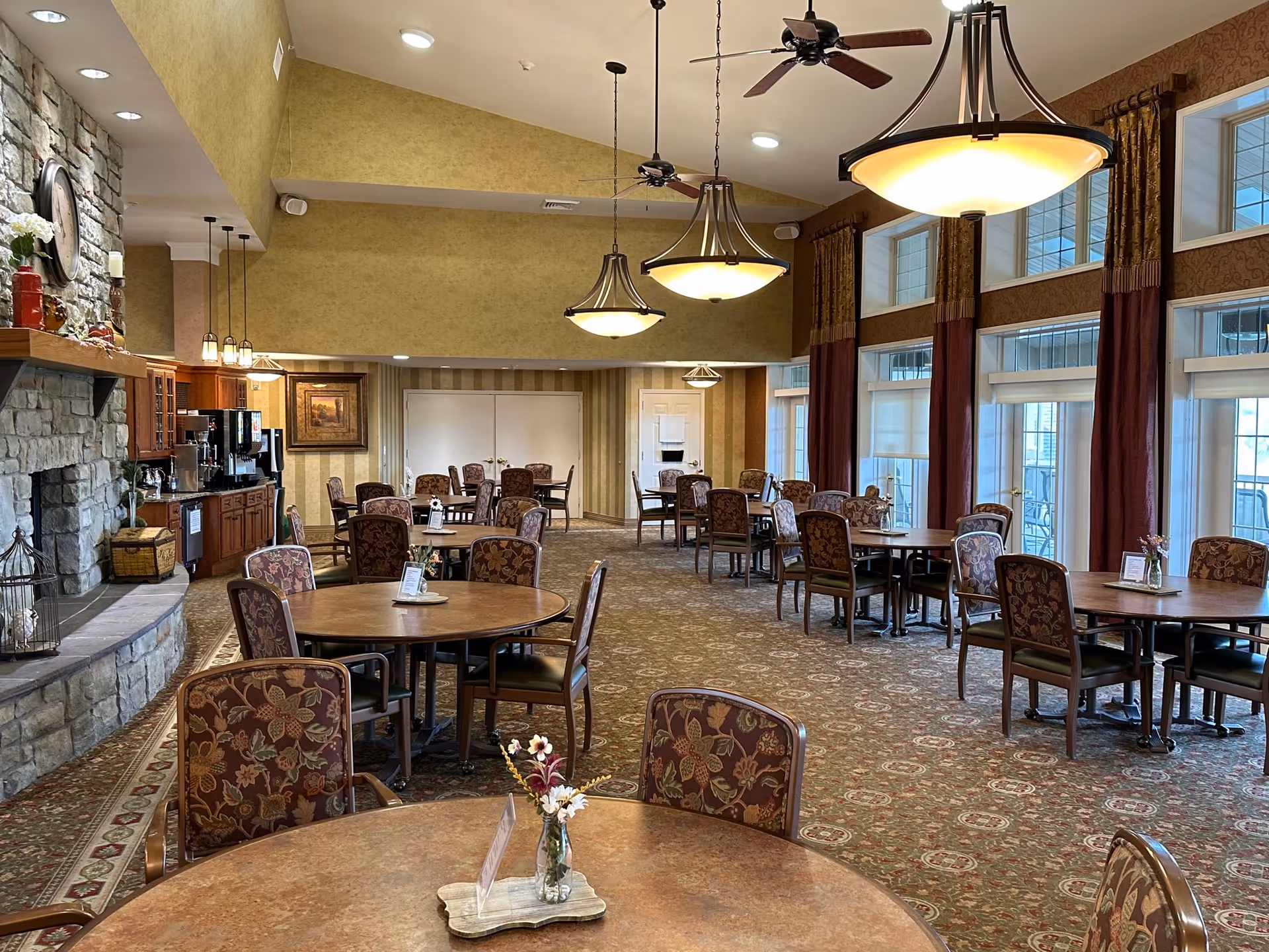 A spacious dining room with multiple round tables and floral upholstered chairs. The room features large windows with maroon curtains, hanging pendant lights, ceiling fans, and a stone fireplace on the left side. There is a coffee and beverage station along the left wall.