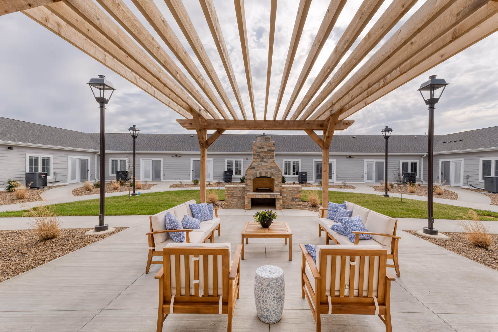 Outdoor seating area at Cedarhurst Senior Living of Owensboro featuring wooden chairs and sofas with blue and white cushions arranged around a wooden coffee table under a wooden pergola. A stone fireplace is centered in the background with a building and lamp posts surrounding the courtyard.