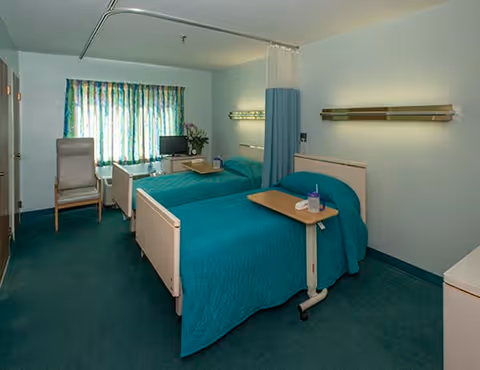 A room with two hospital-style beds covered with teal bedspreads, a small overbed table with a cup and tissue box, a chair near a window with patterned curtains, a small TV on a stand, and a privacy curtain between the beds.