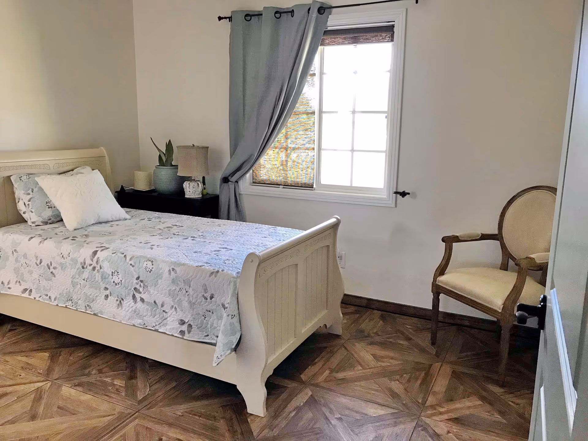 A simple bedroom with a white wooden bed featuring a floral bedspread and two pillows. Next to the bed is a black nightstand with a lamp and a potted plant. A window with gray curtains and a woven blind lets in natural light. There is a wooden chair with a cushioned seat and backrest near the corner of the room. The floor has a patterned wood design.