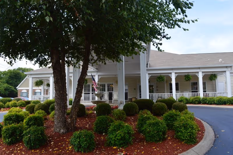 Front exterior view of a single-story building with a covered porch supported by white columns, surrounded by neatly trimmed bushes and a tree in the foreground. An American flag is displayed near the entrance.