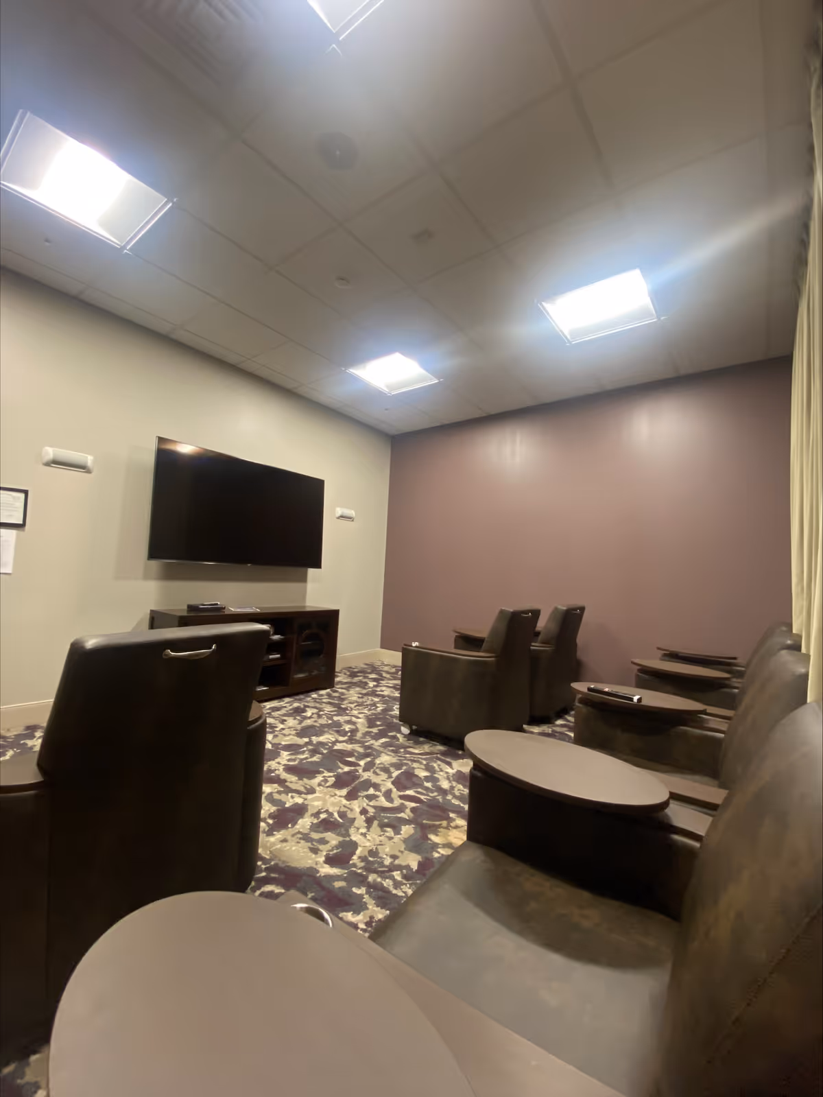 A small theater-style room with several dark leather recliner chairs arranged in rows facing a large flat-screen TV mounted on a light-colored wall. The room has a patterned carpet and a purple accent wall, with bright ceiling lights providing illumination.