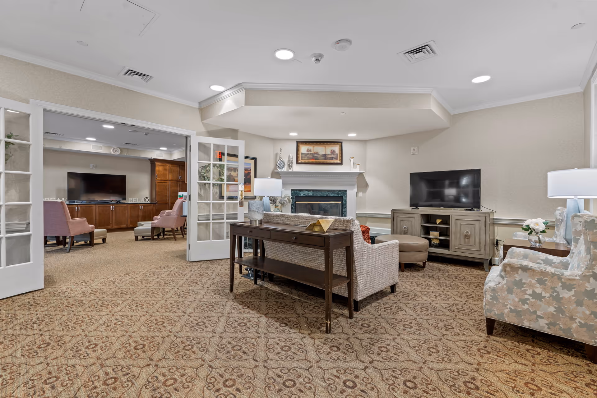 A cozy living room area in a senior living facility featuring a patterned carpet, a fireplace with a framed picture above it, a TV on a wooden cabinet, and comfortable seating including armchairs and a sofa. French doors open to an adjacent room with additional seating and a large TV.