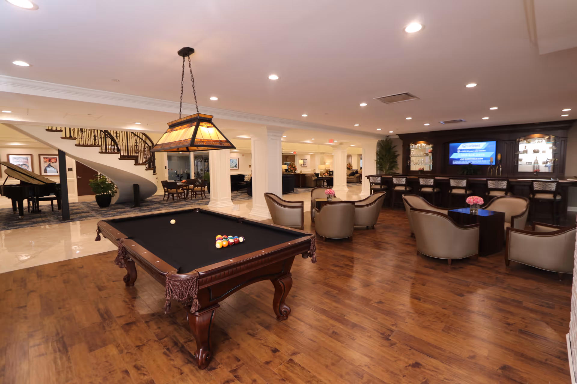 A spacious and well-lit recreational room featuring a pool table with balls arranged for a game, a seating area with several armchairs and small tables adorned with pink flowers, a bar with stools, a large flat-screen TV, and a grand piano near a curved staircase in the background.