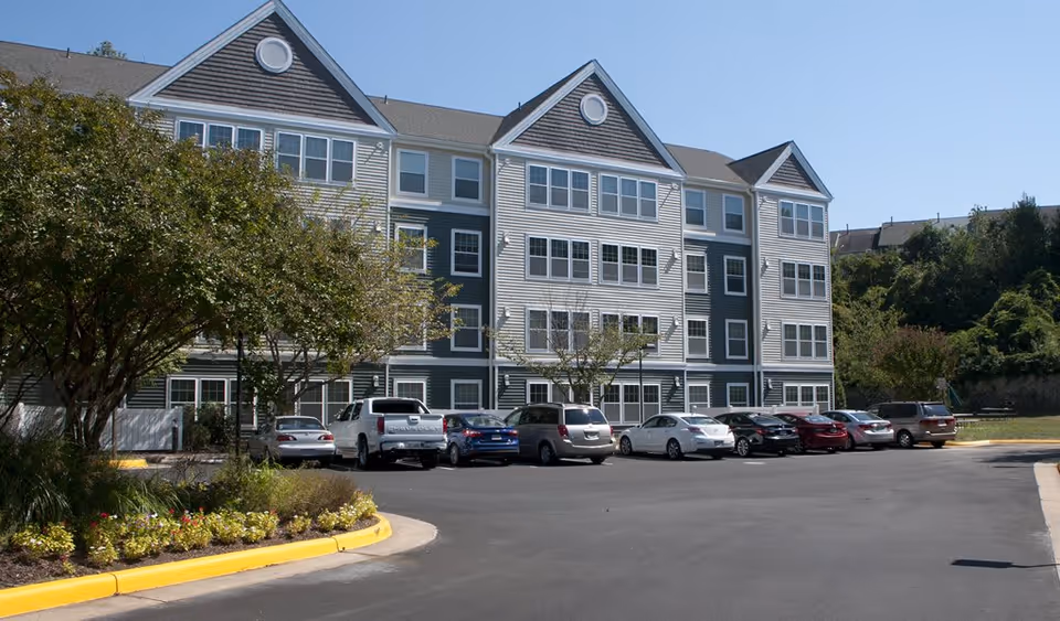 Exterior view of a multi-story senior living facility building with multiple windows, a parking lot with several parked cars, and landscaped greenery including trees and flower beds under a clear blue sky.