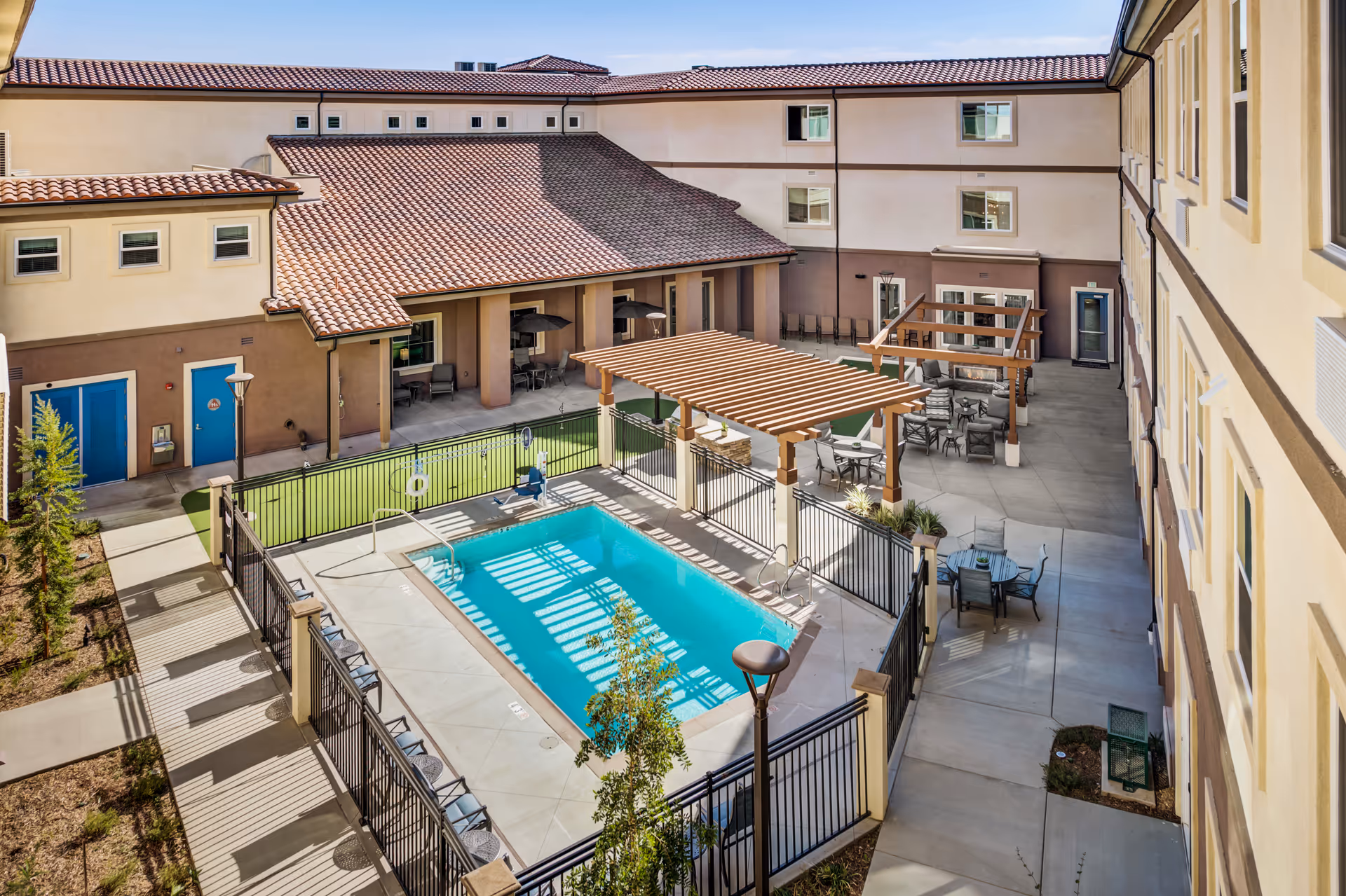 Outdoor courtyard area of WellQuest of Menifee Lakes featuring a fenced swimming pool, several seating areas with tables and chairs under wooden pergolas, and a putting green. The courtyard is surrounded by a multi-story building with beige and brown walls and blue doors.