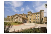 Exterior view of a large, multi-story senior living facility with a stone and stucco facade, arched entrance, and a circular driveway surrounded by landscaping under a partly cloudy sky.