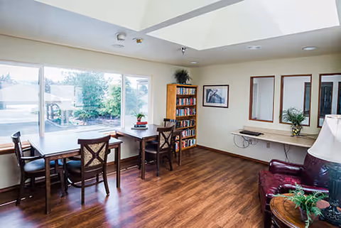 A bright room with large windows showing an outdoor view, featuring a wooden floor, a table with four chairs near the window, a bookshelf filled with books, a wall-mounted desk with a plant, a framed picture on the wall, and a leather armchair with a side table and lamp.