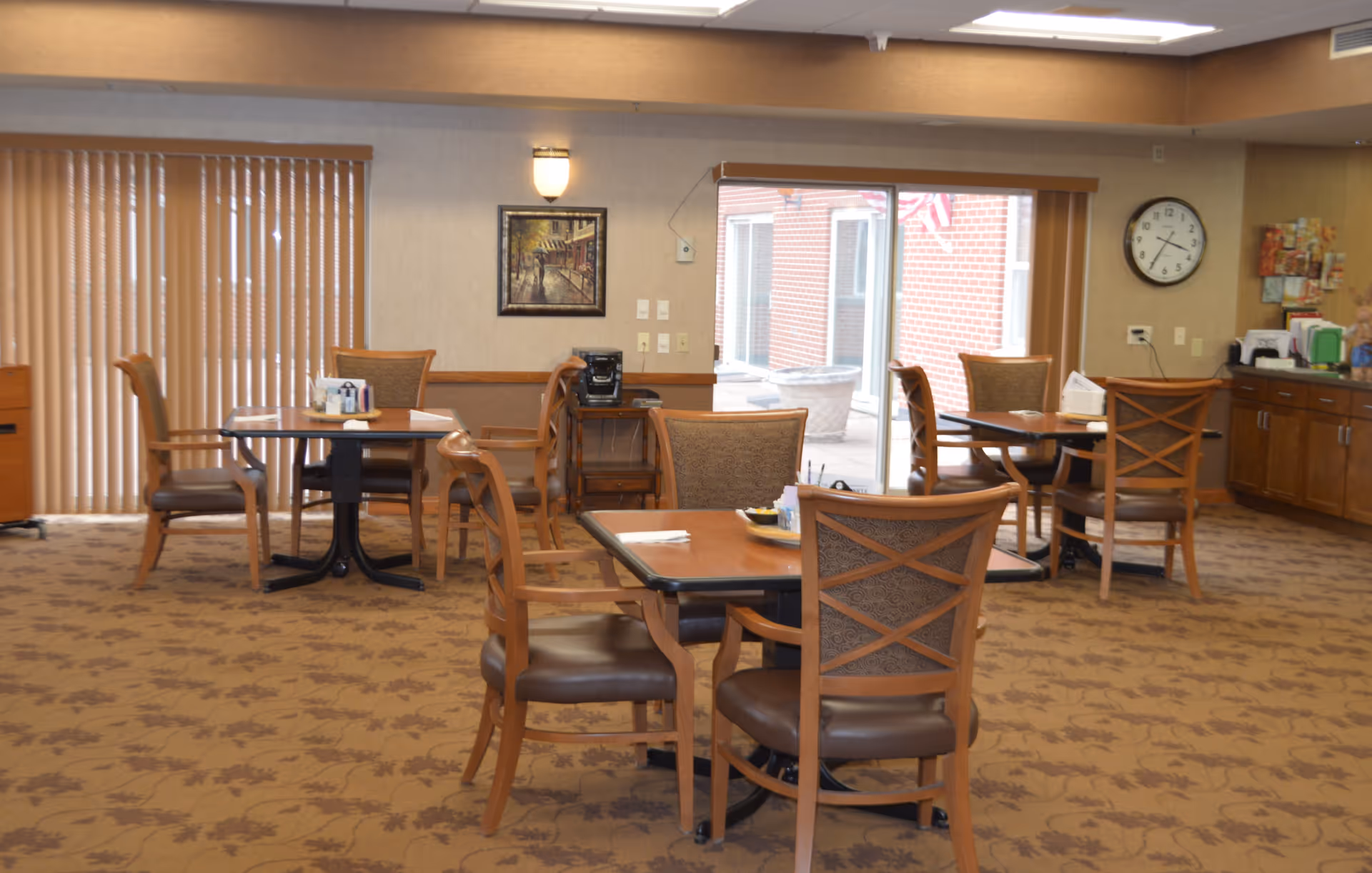 A dining area in an assisted living facility with several wooden tables and chairs arranged on a patterned carpet. The room has large windows with vertical blinds, a wall clock, a painting, and a counter with various items on it. The space is well-lit with ceiling lights and a wall sconce.