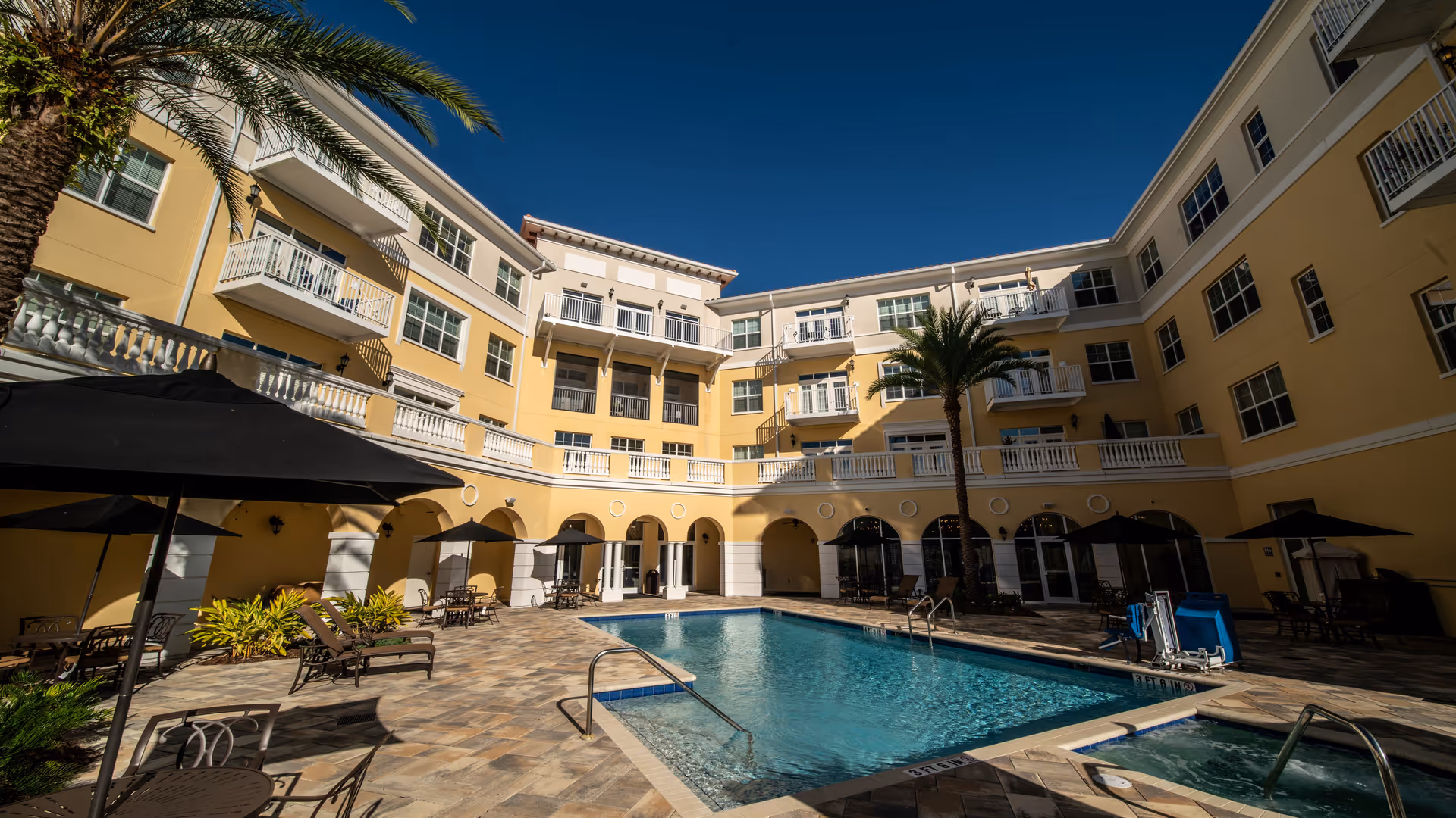 Outdoor courtyard area of a senior living facility with a swimming pool and hot tub surrounded by lounge chairs, tables with umbrellas, palm trees, and a three-story yellow building with balconies under a clear blue sky.