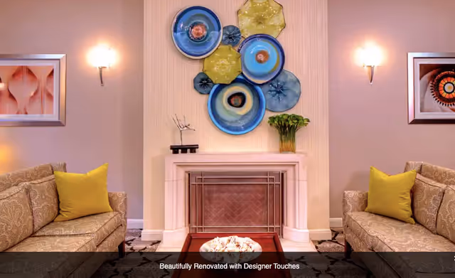 A cozy living room with two patterned beige sofas facing each other, each with a yellow cushion. Between them is a wooden coffee table with a decorative bowl. The room features a white fireplace with a metal screen, above which hangs a colorful wall art installation of circular shapes in blue, yellow, and black. On either side of the fireplace are framed abstract artworks and wall sconces providing warm lighting.