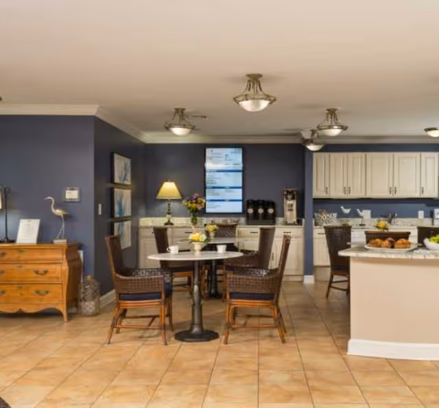 Communal dining area with round tables and wicker chairs in front of a kitchenette with white cabinets, a coffee station, and decorative accents.