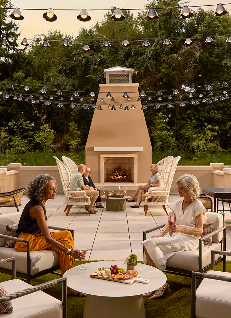 An outdoor patio area with a large beige fireplace in the center. Four older adults are seated in comfortable chairs around small tables, enjoying drinks and snacks. String lights hang overhead, and there is greenery and trees in the background.