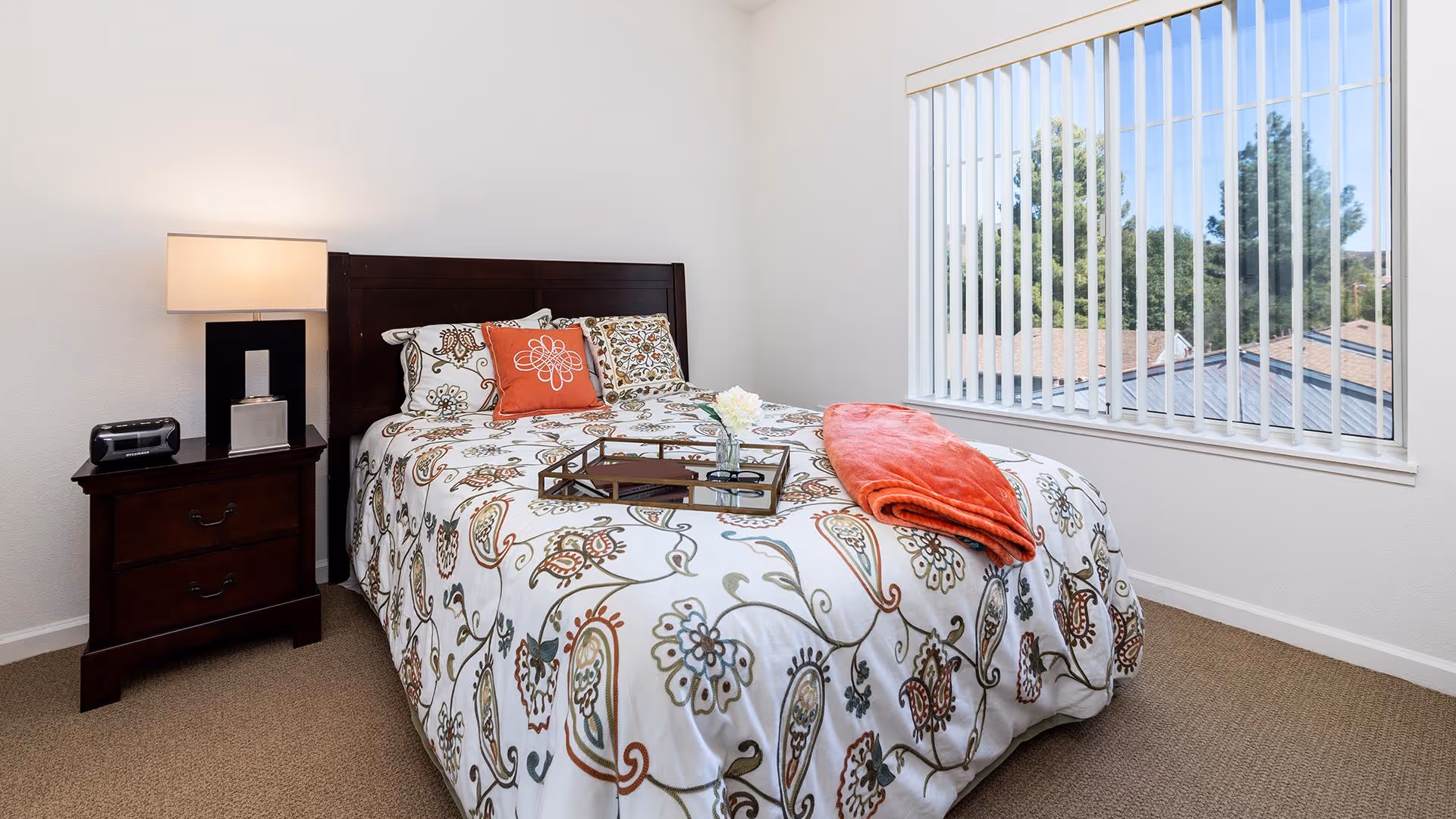 A bedroom with a bed featuring a floral patterned comforter and decorative pillows, a wooden headboard, a nightstand with a lamp and a clock radio, and a window with vertical blinds showing trees and rooftops outside.