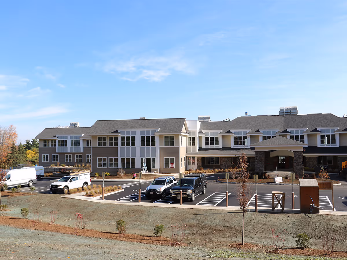 Exterior view of a two-story senior living facility building with a parking lot in front. Several vehicles are parked, and the building features large windows and a covered entrance. The sky is clear and blue.