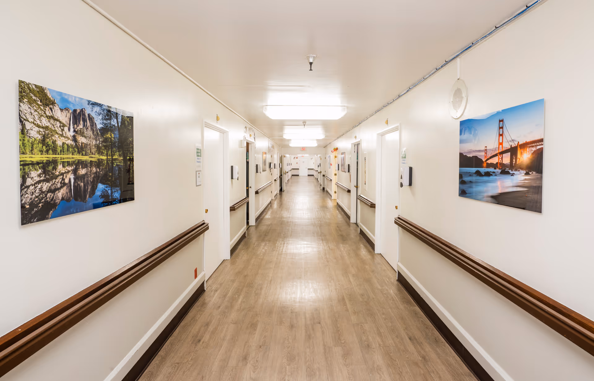 Long, well-lit hallway in a senior living facility with handrails and framed landscape photos on the walls.