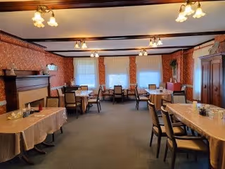 A dining room with several tables covered with beige tablecloths and chairs arranged around them. The room features patterned wallpaper, multiple windows with white curtains, a fireplace on the left side, and warm ceiling lights. There is a wooden cabinet on the right side and some decorative plants.