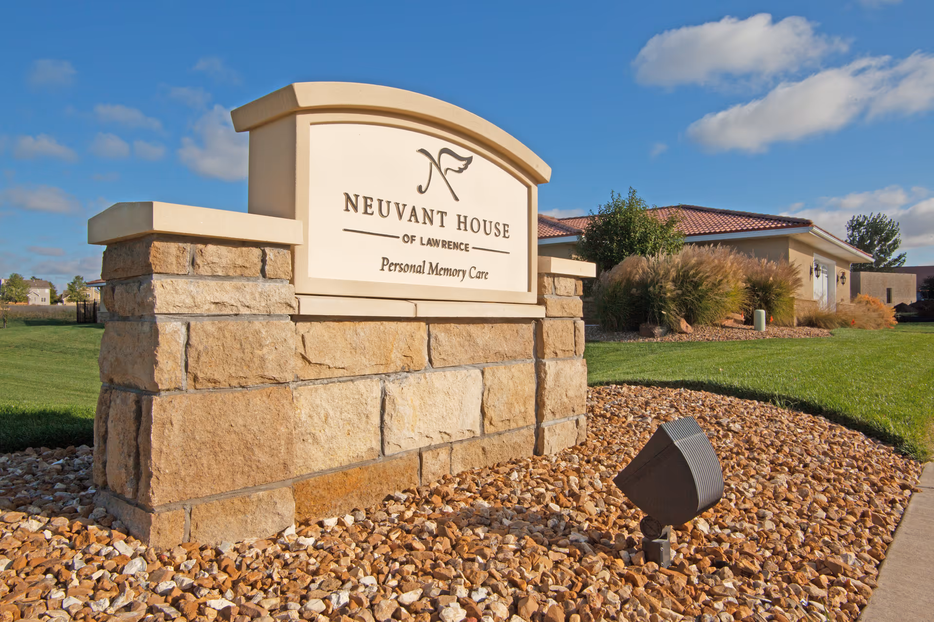 Stone sign for Neuvant House of Lawrence Personal Memory Care with a landscaped area of rocks and grass, and a building with a red-tiled roof in the background under a partly cloudy blue sky.