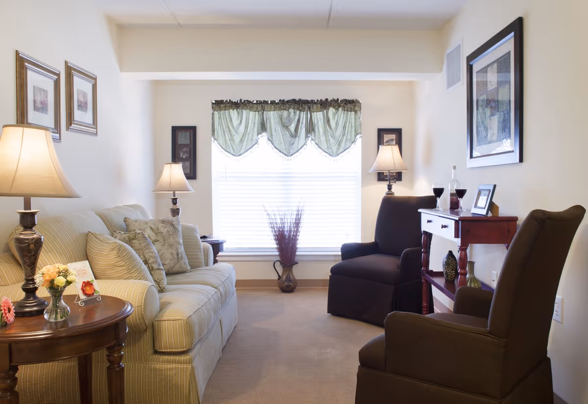 A cozy living room with a beige striped sofa adorned with floral cushions, two dark brown armchairs, and a wooden side table with a lamp, flowers, and a welcome card. A window with green valance curtains is centered on the back wall, flanked by two framed pictures and two table lamps. A small wooden console table holds a bottle of wine, two glasses, and decorative items.