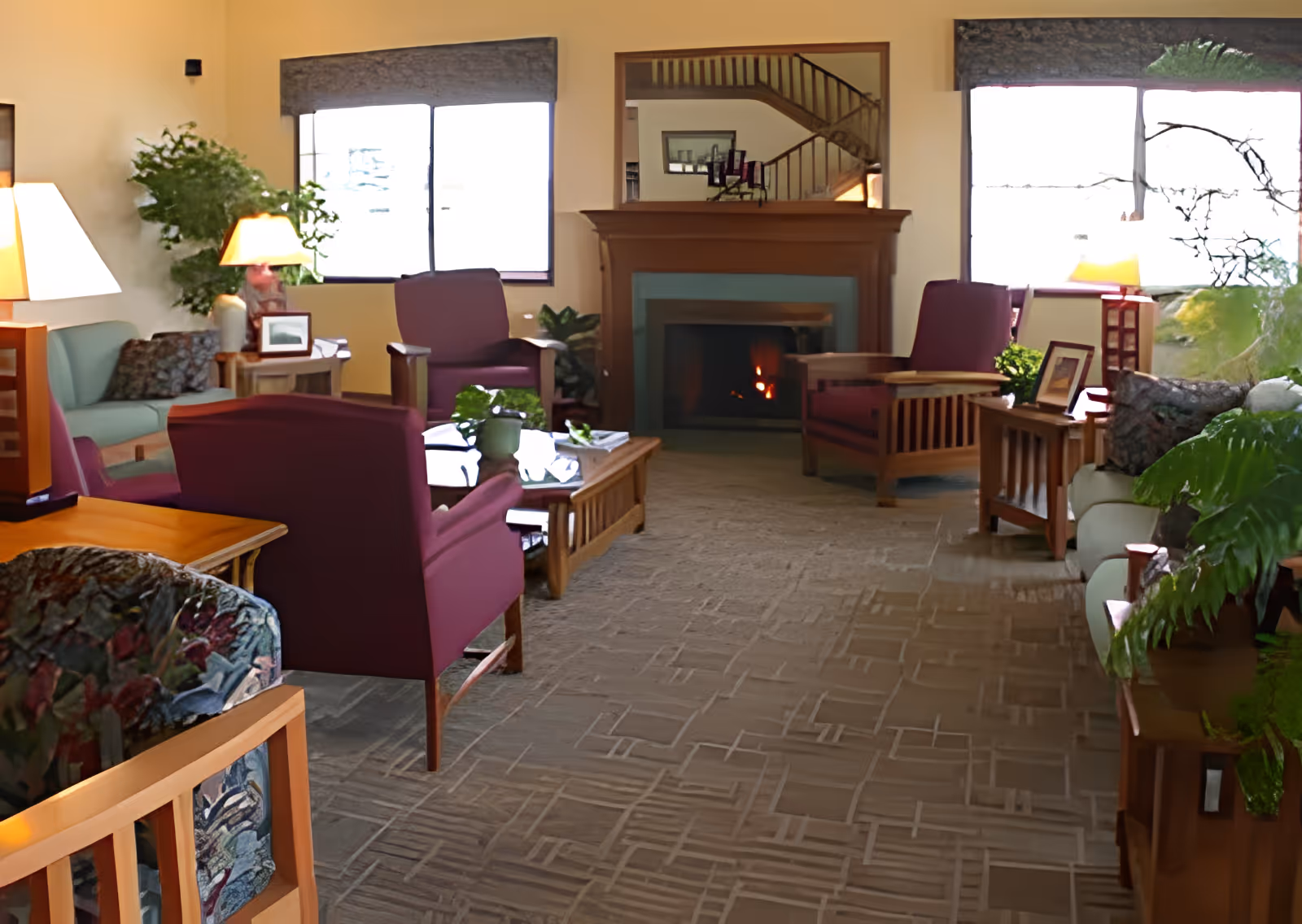 A cozy living room area in an assisted living facility with multiple armchairs and sofas arranged around a wooden coffee table. There is a fireplace with a fire burning, a large mirror above it reflecting a staircase, and large windows letting in natural light. The room is decorated with plants, table lamps, and framed pictures.