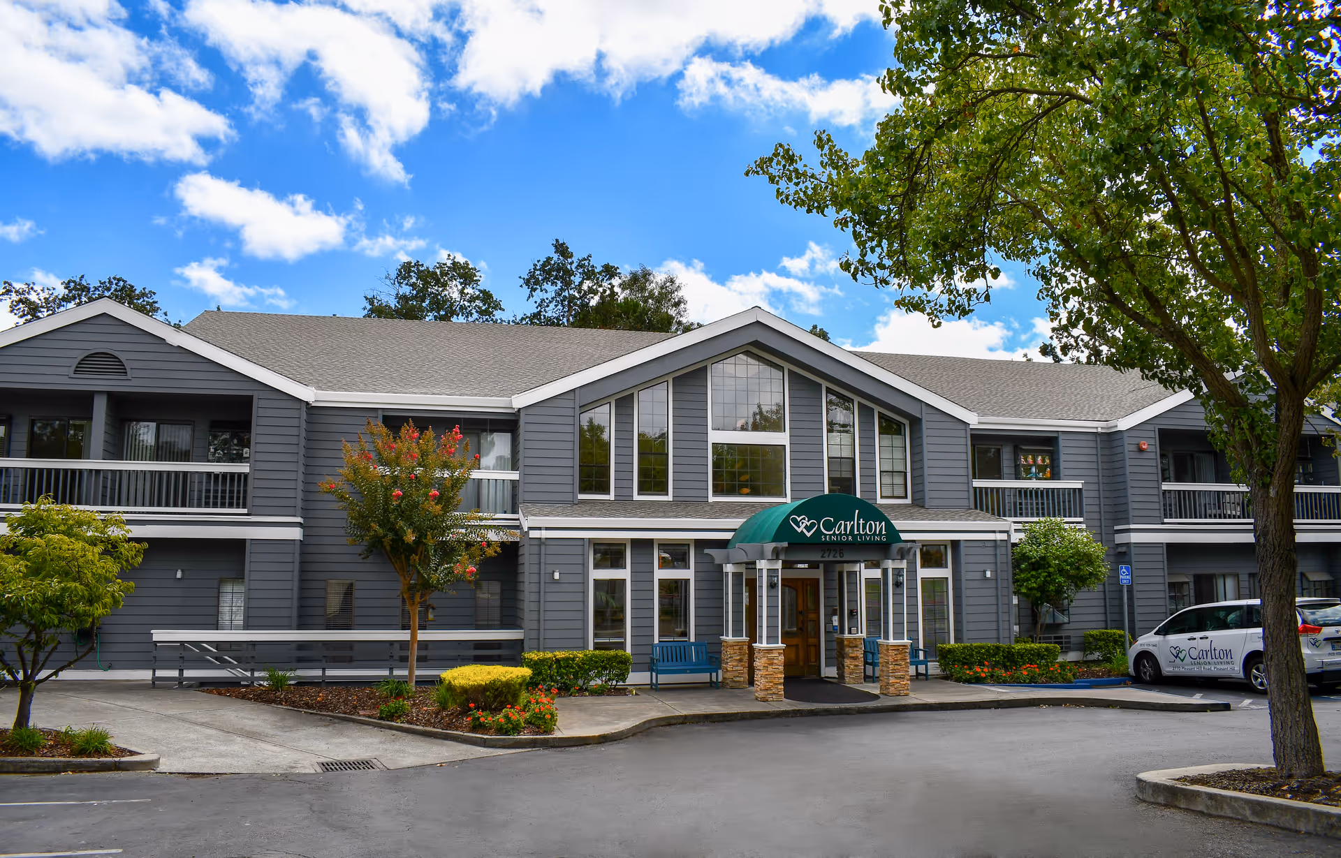 Front exterior of a two-story gray Carlton Senior Living building with a green awning over the main entrance, landscaping and a parked car.