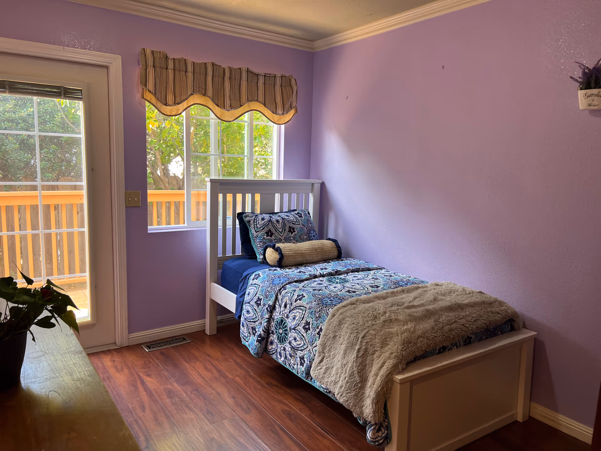 A small bedroom with lavender walls and wooden flooring. There is a single white bed with a blue and white patterned bedspread, a cylindrical pillow, and a beige throw blanket. A window with a striped valance and a glass door open to an outdoor wooden deck. A plant is partially visible on a wooden surface in the foreground.