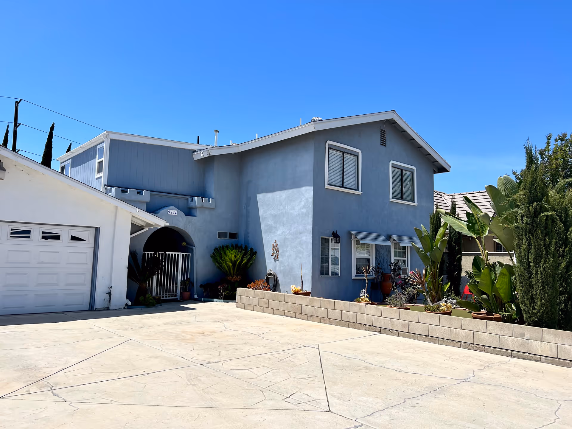 Exterior view of a two-story residential building painted blue with white trim, featuring several windows with blinds, a small garden area with various plants, and a concrete driveway in front. The sky is clear and blue.