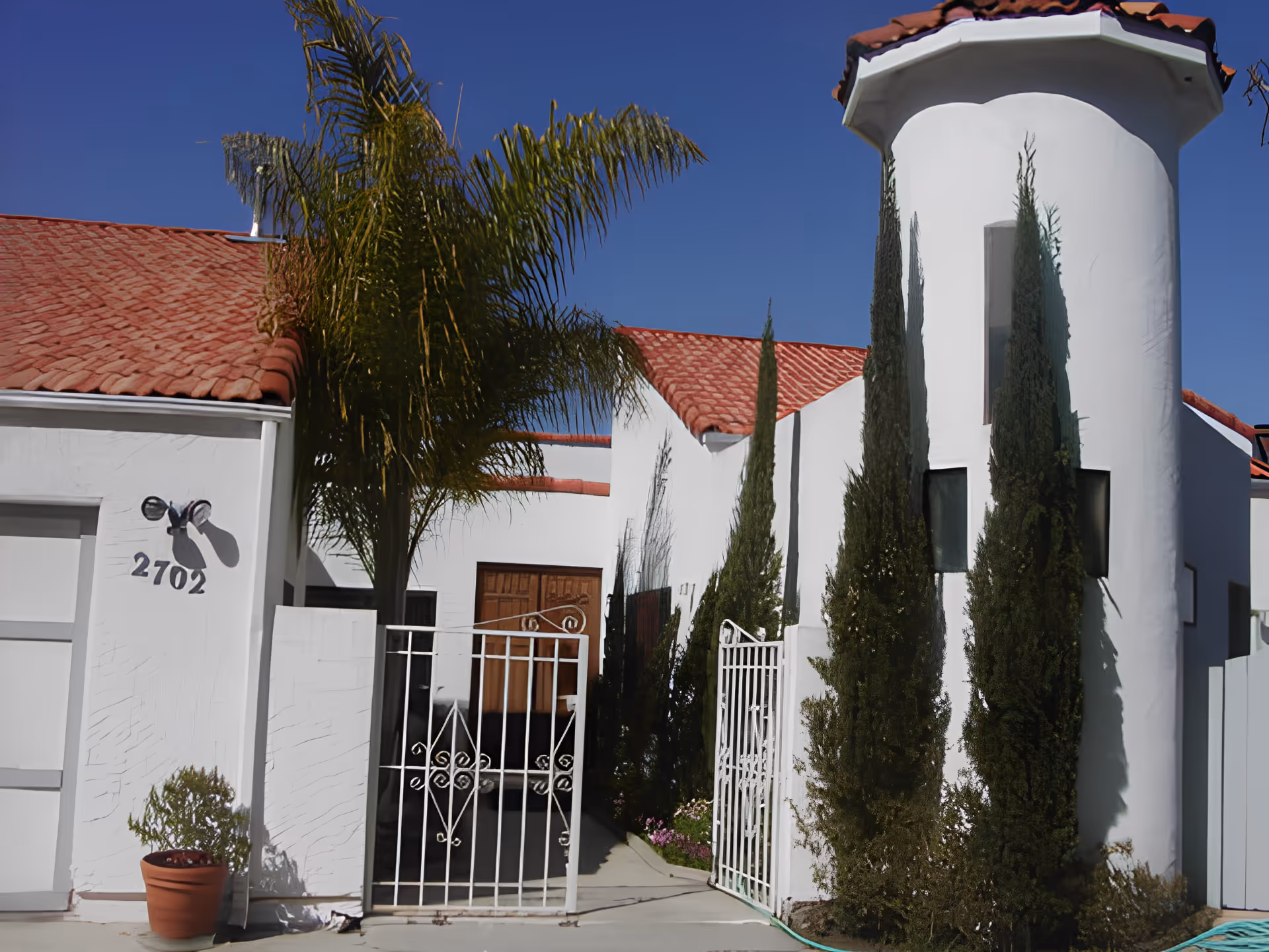 White stucco building with a red tile roof and gated entrance flanked by palm and cypress trees, showing the address 2702.