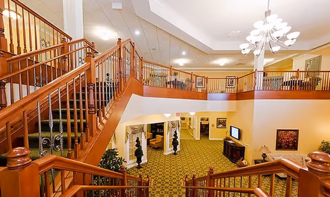 Two-level atrium lobby with a central wooden staircase, patterned carpet, balcony railings, seating areas, and a chandelier.