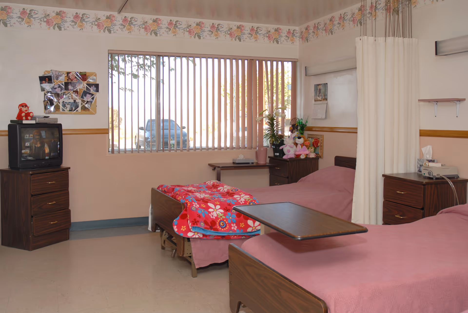 Two hospital-style beds with pink bedding in a convalescent facility room with nightstands, a TV, and a window with vertical blinds.