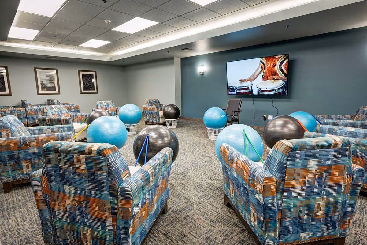 A room with patterned armchairs arranged in a circle around exercise balls placed on white baskets, with drumsticks resting on some balls. A flat-screen TV mounted on a blue wall displays a person playing drums. The room has a carpeted floor, framed artwork on the walls, and ceiling lights.