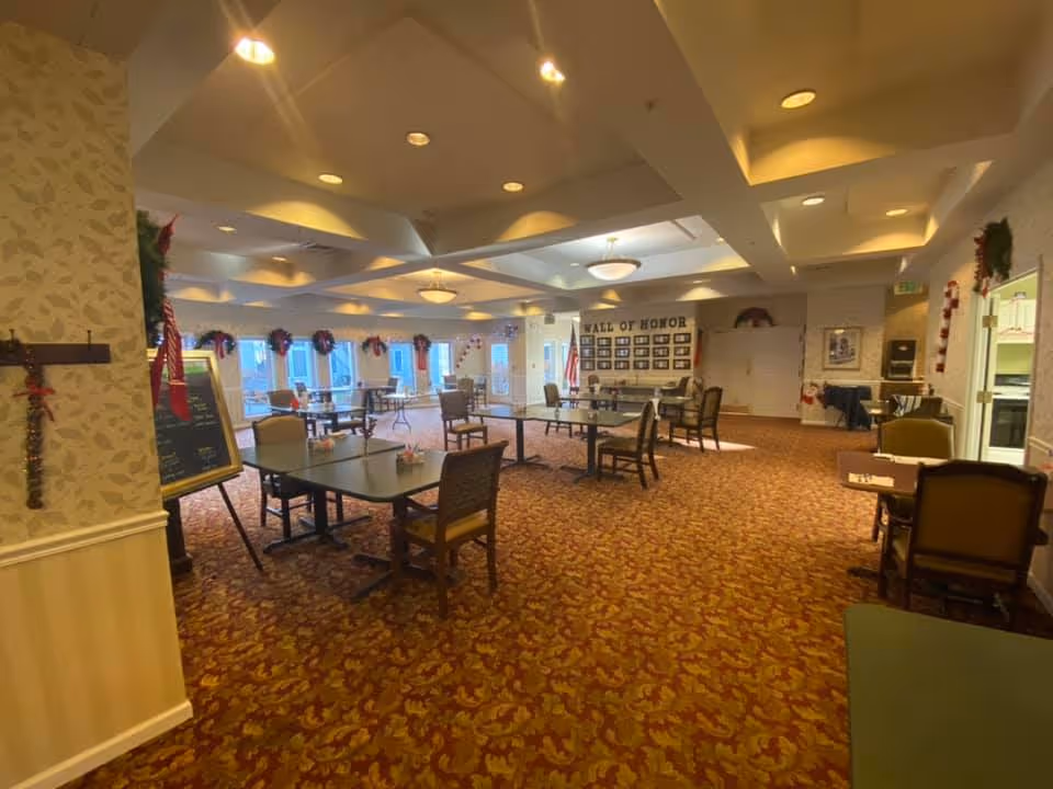 A spacious dining room with multiple tables and chairs arranged on a patterned carpet. The room is decorated with holiday garlands and wreaths along the windows and walls. There is a 'Wall of Honor' display on the far wall, and the ceiling has recessed lighting and decorative light fixtures. The room has a warm and inviting atmosphere.