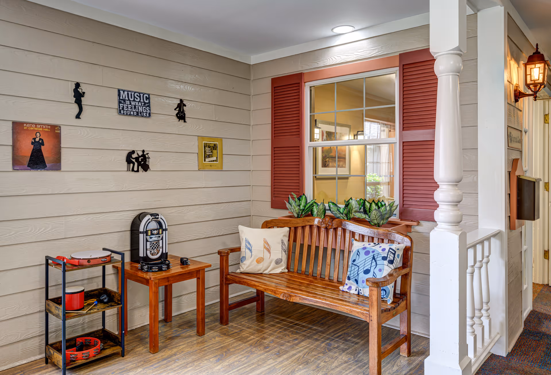 Wooden bench with musical-themed pillows, a small table holding a jukebox and shelving with instruments against a shiplap wall decorated with music-themed wall art and a window with red shutters.