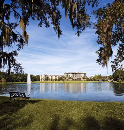 A peaceful outdoor scene at Lake Port Square featuring a large pond with a water fountain in the center, a bench on the grassy shore, and a multi-story building in the background under a blue sky with scattered clouds. Large trees with hanging moss frame the top of the image.